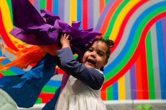 Little girl standing in front of brightly coloured wall painting waving a bundle of brightly coloured crepe paper
