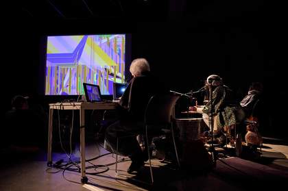 Three people seated on a stage, lit by spotlights, behind desks with audio equipment and laptops sitting on them. In front of them, there is a screen displaying an abstract purple and yellow image.