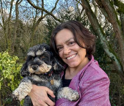 A woman standing in a woodland smiling and holding a dog