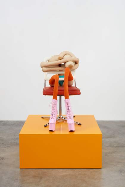 A form with breasts sits upon a red and white chair upon a bright orange cube.