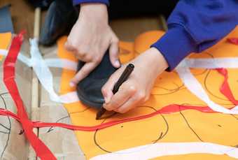 A young person drawing with a black pen onto orange paper covered in red and white tape.