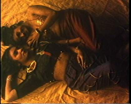 Two women of colour lie on yellow fabric, their bodies touching each other, one woman looks ahead while the other, touching her own shoulder smiles at her companion.