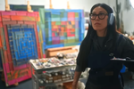 Zadie Xa stares at a canvas off camera. She holds a palette and wears headphones. Behind her are many abstract paintings made of colourful squares.
