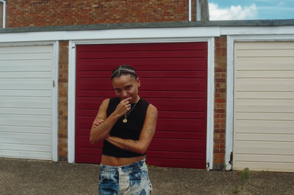 Rene Matić stands in front of a row of garages or storage spaces. The door on the left is white, the centre door is red, and the door on the right is cream. They have their arms crossed and one hand on their chin, giving the camera a slight smile.