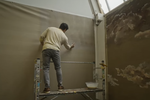 Mohammed Sami stands on scaffolding, priming a large piece of linen which will become a canvas. His back is to the camera.