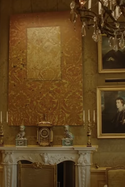 An installation shot of one of Mohammed Sami's paintings over an ornate white marble fire place. The painting features yellow brocade wallpaper with a lighter patch in the centre, suggesting a painting or picture has been removed from the wall.