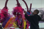 Nnena Kalu ties pieces of fabric around a large, multi-coloured sculpture. She is wearing a black hoodie and gold hoop earrings. Behind her are two of her vortex drawings.