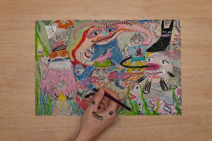 A piece of paper on a table filled with drawings in lots of different colours. At the bottom of the picture is a hand holding a pencil and drawing onto the paper. The hand has eyes and a mouth drawn onto it, smiling up at the viewer.