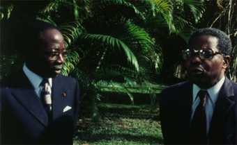 Two men in suits against a backdrop of vegetation