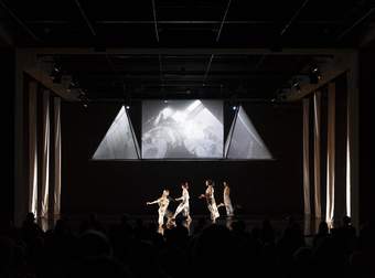 A dark image with four people lit up and behind them are three screens. Either side are four strips of fabric draped from ceiling to the floor.