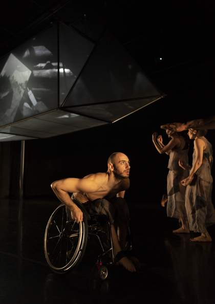 Dancer in a wheelchair with two dancers wearing natural coloured clothes standing to the side.