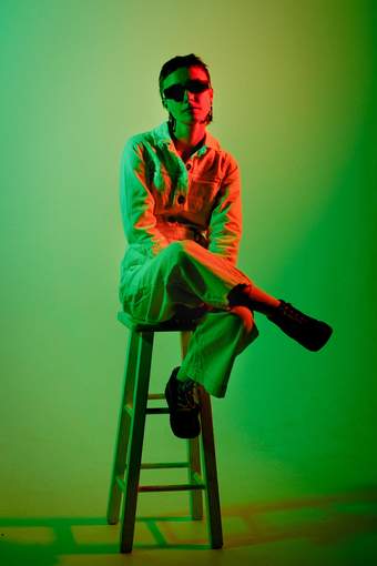 A photograph of a person sat on a stool with a green background.