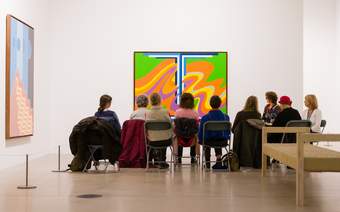 A group of visitors listening to a talk by Tate specialist Susanna Webster, who is sitting to the right, in front of a large colourful artwork