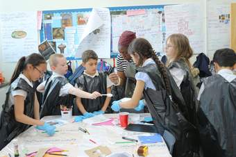 This is a photograph of a group of school children with artist Victoria. Victoria is wearing a black and white striped top, a grey apron and red and white headscarf. She is leaning over a table, holding a plastic fork in one hand, with a roll of tape on h