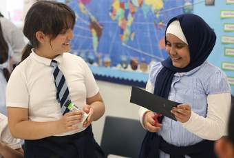 A photograph of two children participating in craft workshops at a school. The girl on the left is holding a glue stick and the girl on the right is using a pair of scissors.