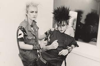 Two punk women one leaning against a window wit a broken fragment of mirror on the winder. One woman is blonde and the other has dark hair which is spiked up and they are wearing heavy make up, denim and metal jewellery.