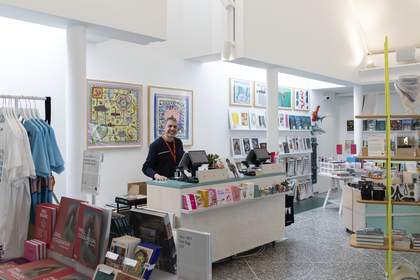 A colourful shop filled with books, artworks, t-shirts and other items on display. A smiling Shop Assistant stands behind the till.