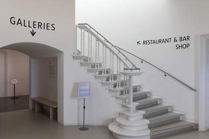 A staircase inside the gallery, with a sign pointing up towards the restaurant, bar and shop. To the left of the staircase, a sign points forward reading ‘Galleries’.