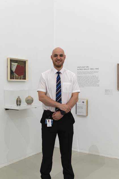 A smiling Security Officer wearing a shirt and tie, standing in front of a gallery wall displaying artwork by Alfred Wallis.