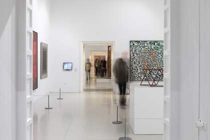 A room in the gallery with white walls and artworks hanging on the walls. There is a large white plinth with an artwork standing on it, and cord barriers surrounding the art.