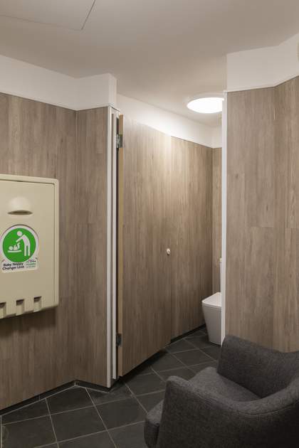 The interior of the parent and baby room, with a toilet cubicle, nappy changer unit and grey armchair.