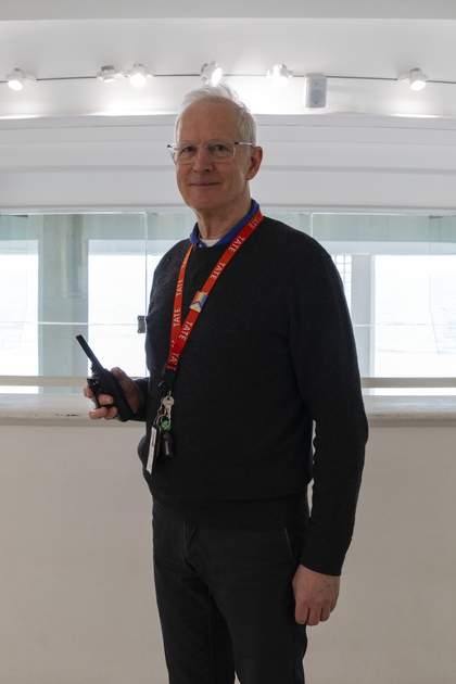 A smiling Visitor Assistant wearing an orange Tate lanyard decorated with an LGBTQ+ badge, holding a walkie talkie.