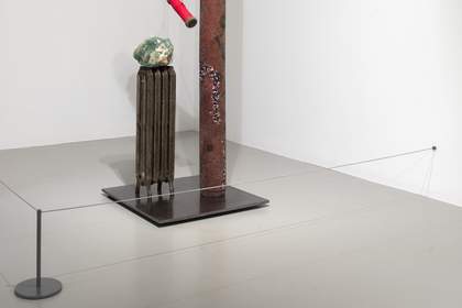 A metal sculpture standing on the floor, surrounded by a thin metal cord barrier.