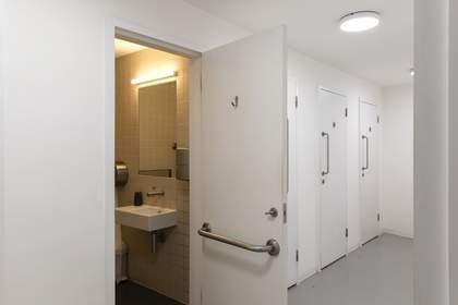 A series of four toilet cubicles with one door propped open, showing a mirror, sink and handrail.