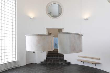 A curved room in the gallery with a large, curved, white paper artwork suspended from the ceiling. There are stairs leading into another room.