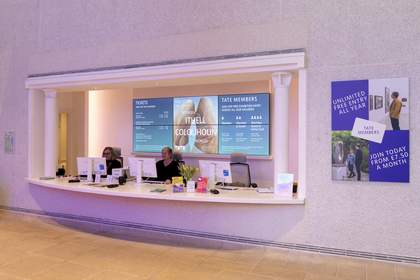 The ticketing desk in the lobby. Two staff members sit behind the desk. Behind them there is a large screen with ticketing and membership information displayed on it.