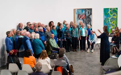 A large community choir stands in a semi-circle inside a curved art gallery, singing while a conductor leads from the foreground