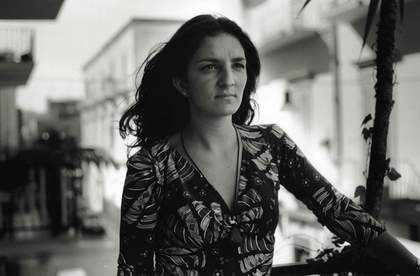 A black and white image showing a white woman with long dark hair, she wears a patterned dress with a V neck and stands in a street.