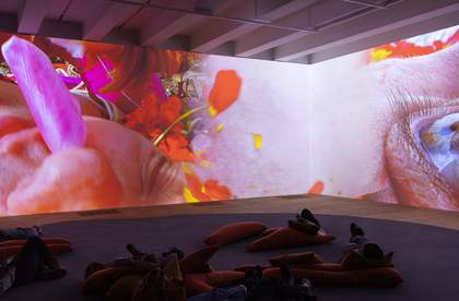 Corner of a large room, carpet on the floor with people lying on bean bags, large screen with image of extreme close up of an eye and a face.