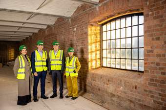 A group of four people wearing high-vis jackets and hard hats are standing next to the left of a window in a building site.