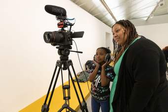 A photograph of a child wearing headphones standing next to an artist looking at a camera screen