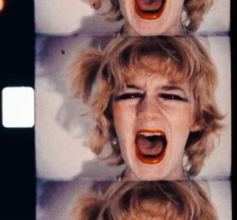 A film still of a woman looking directly down the barrel of the camera with her mouth open wide screaming