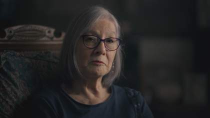 'Close up shot of an older mixed British and Chinese woman, with grey hair and glasses, wearing a navy blue top.