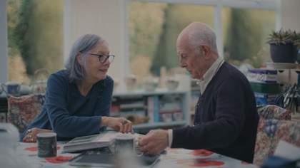 An older couple sit at a table, looking at a family photo album.