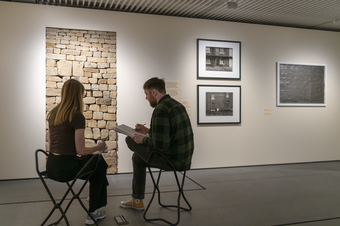 A photograph of two visitors sat on portable chairs in a gallery space at Tate Liverpool + RIBA North sketching in front of a brick sculpture