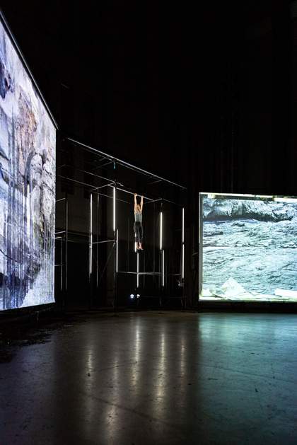 Dancer hanging from a pole in the ceiling of the stage in front of light strips and with a large screen either side