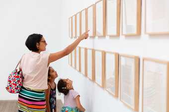 a parent pointing at an artwork on the wall with two children looking up positioned in front of her.