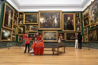 A person in a red dress with children sits on a bench in a salon hang gallery looking at historial paintings