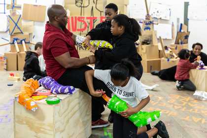 An adult at a worskhop with some children who are making clothes for him out of scrap.