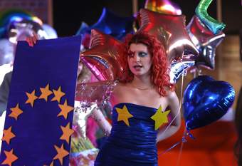 A photograph of a performer at Tate Liverpool wearing a blue dress with two yellow stars on the front. She is carrying a Euro flag and there are lots of balloons in the background.
