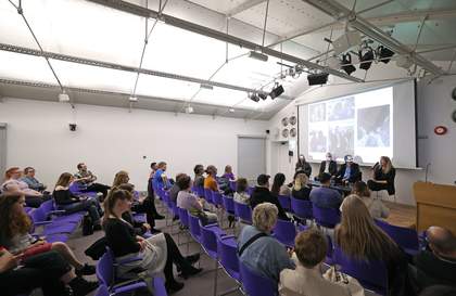 A photograph of people sat in the auditorium listening to a panel of people on a small stage at the front. There is also a screen behind the panelists with pictures projected on it.