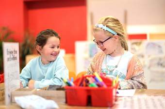 Two young children are sat colouring at a table, both smiling