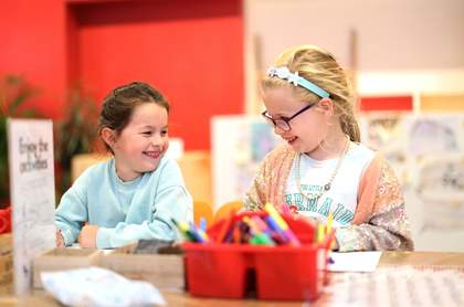 Two young children are sat colouring at a table, both smiling