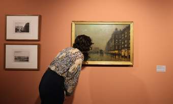 A photograph of a visitor looking closely at a painting display on an orange wall.