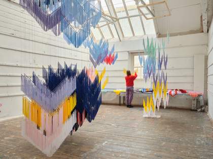 The artist is standing towards the top of a ladder in her studio in St Ives, hanging elements of an artwork made of threads from frames suspended from the ceiling. The studio is lit by natural light from the skylights above.