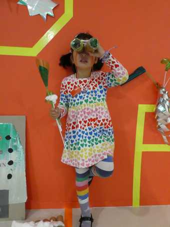 Image shows young girl in bright clothing looking through a pair of space binoculars she has made and holding a space flower she has made in the other hand. She stands against an orange wall with other shiny space props on the wall beside her.